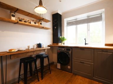 A kitchen with wooden shelves jars cups two black stools and a washing machine under a window at Rosedene Rickinghall