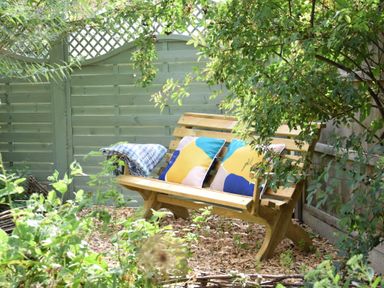An outdoor wooden bench with pillows and a blanket surrounded by plants and a wooden fence at Rosedene Rickinghall
