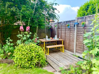 An outdoor garden area with a wooden table and benches on a deck next to a wooden fence with hanging flower pots and string lights at Rosedene Rickinghall
