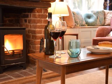 A living room with a wooden table holding a bottle of wine two wine glasses a blue tumbler plates with forks and a loaf of bread by a lit fireplace at Rosedene Rickinghall