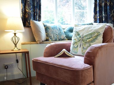 A pink armchair with a butterfly cushion and open book beside a window with patterned curtains and a side table with a lamp at Rosedene Rickinghall