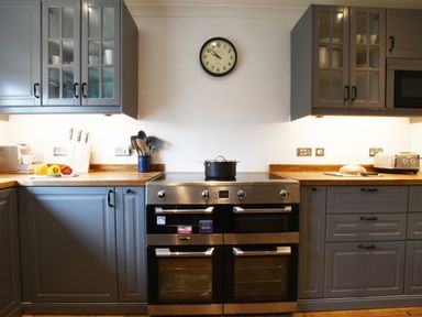 A kitchen with a stove and oven in the center a clock on the wall and gray cabinets with a toaster and knives on the wooden countertops at Rosedene Rickinghall