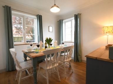 A dining area with a wooden table set with wine glasses and bowls surrounded by white chairs near windows with curtains at Rosedene Rickinghall