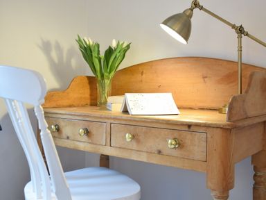 A wooden desk with brass lamp and vase of flowers next to a white chair at Rosedene Rickinghall