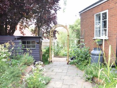 A garden pathway with a wooden archway next to a brick house and a shed at Rosedene in Rickinghall