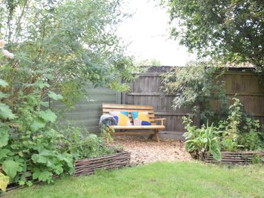 A wooden bench with cushions and a blanket in a garden corner surrounded by plants and a wooden fence