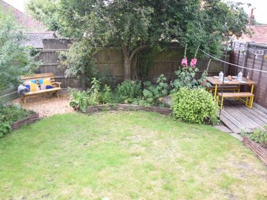 A garden with a wooden bench with cushions a tree a flower bed and a dining table with benches on a wooden deck at Rosedene Rickinghall