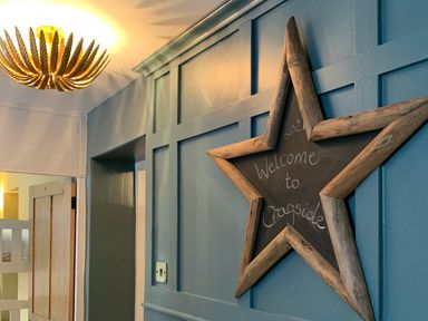 A blue paneled wall with a star-shaped chalkboard sign that says welcome to Cragside with a wooden door and a ceiling light fixture at Cragside Aldeburgh