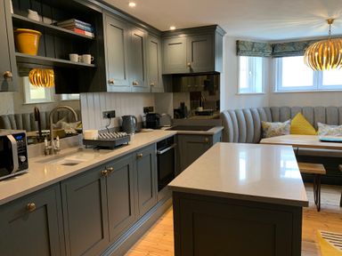 A kitchen with gray cabinets a countertop island a built in oven a curved bench seat and a dining table at Cragside  Aldeburgh