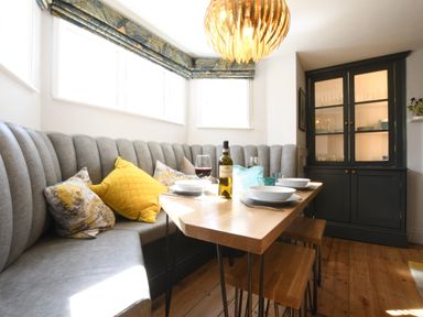 A dining area with a wooden table set with plates and wine glasses next to a curved gray banquette and a dark cabinet at Cragside Aldeburgh