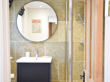 A bathroom with a round mirror above a sink and a shower area behind a wooden door at Cragside Aldeburgh
