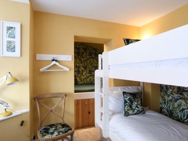 A bedroom with a white bunk bed a wooden chair with a tropical cushion a yellow desk lamp and framed pictures at Cragside  Aldeburgh