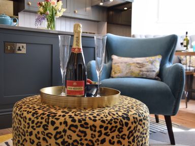 A living area with a bottle of champagne and two glasses on a tray on a leopard print ottoman with a blue armchair in the background at Cragside Aldeburgh