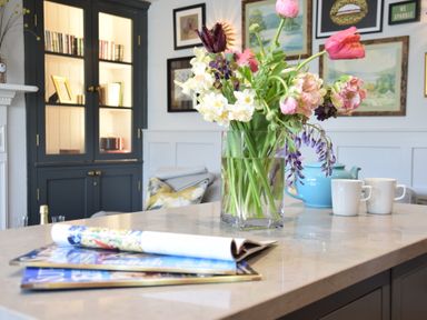 A kitchen island with a vase of flowers, two white mugs, a blue teapot, and magazines with a cabinet and framed pictures in the background at Cragside, Aldeburgh