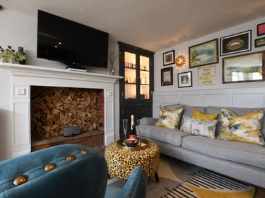 A living room with a grey sofa and patterned cushions a leopard print ottoman a blue armchair stacked firewood in a fireplace and framed pictures on the wall at Cragside Aldeburgh