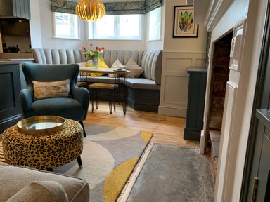 A seating area with a blue armchair a patterned ottoman a corner bench with cushions a dining table and a fireplace at Cragside Aldeburgh