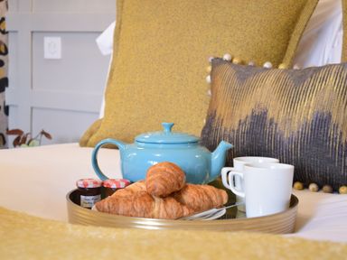 A tray with a blue teapot two white cups croissants and jam jars on a bed with yellow pillows at Cragside  Aldeburgh