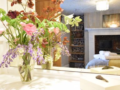 A flower arrangement in a glass vase on a white surface with a mirror reflecting a room with shelves folded towels and a fireplace at Cragside Aldeburgh