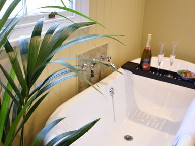 A bathroom with a white bathtub near a window and a green plant with a bottle of champagne and two glasses on the bathtub tray at Cragside Aldeburgh in Aldeburgh