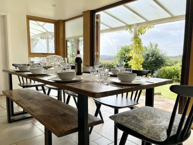 A dining area with a wooden table set with bowls glasses and a bottle and black chairs and a bench near open glass doors at Skelder Parham in Parham Near Framlingham