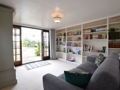 A room with a gray sofa and built in bookshelves next to sliding glass doors opening to a garden at Skelder Parham in Parham Near Framlingham
