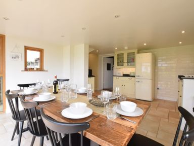 A dining area with a wooden table set with plates bowls glasses and cutlery next to a kitchen with cream cabinets and a fridge at Skelder Parham in Parham Near Framlingham