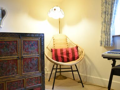 A corner with a woven chair with a striped pillow a floor lamp an ornate cabinet and a keyboard at Skelder Parham in Parham Near Framlingham