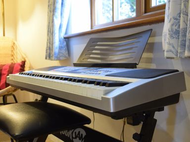 A keyboard piano with a black bench near a window with blue patterned curtains at Skelder Parham in Parham Near Framlingham