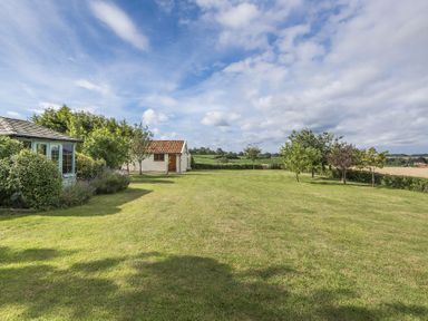 A large grassy garden with a small white building and trees under a partly cloudy sky at Skelder Parham in Parham Near Framlingham