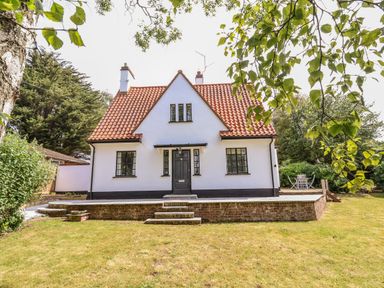 A white house with a red tiled roof a black door and windows surrounded by a garden with grass and trees at High Ground Reydon