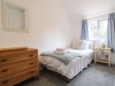 A bedroom with a single bed, wooden chest of drawers, bedside table with lamp and window with curtains at High Ground, Reydon