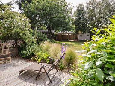 A garden with a reclining chair on a wooden deck and a wooden bench beside plants and trees at High Ground Reydon