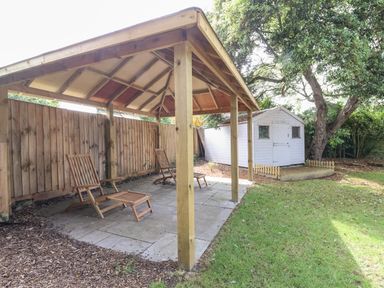 A garden area with a wooden gazebo containing two wooden chairs and a white shed beside a large tree at High Ground Reydon in Reydon