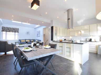 A dining table set with plates and wine glasses next to a kitchen with white cabinets and a stainless steel range hood at Pier View Southwold