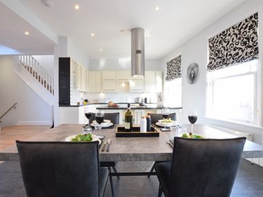 A dining table set with plates glasses and wine in a kitchen with white cabinets and patterned window blinds at Pier View Southwold