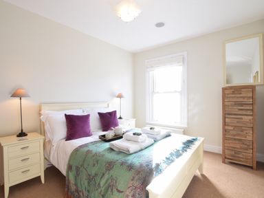 A bedroom with a bed with purple pillows and a green blanket two bedside tables with lamps and a wooden chest of drawers with a mirror at Pier View Southwold in Southwold