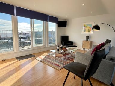 A living room with large windows overlooking a balcony with chairs a glass coffee table and seating at Pier View Southwold