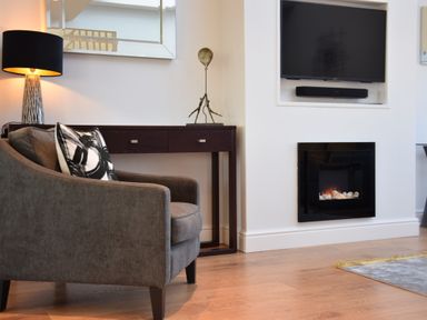 A living room with a gray armchair a black and white pillow a table lamp a console table with a sculpture a built in black fireplace and a mounted television at Pier View Southwold