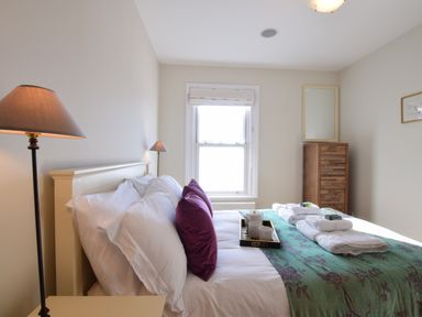 A bedroom with a bed with white and purple pillows a green bed cover and towels on top a wooden cabinet a floor lamp and a window at Pier View Southwold in Southwold