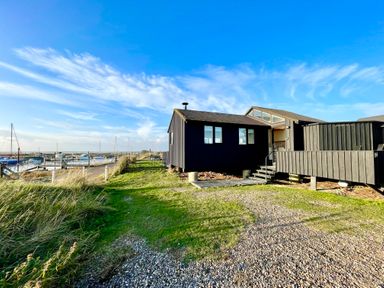 A black wooden building near a dock with boats under a blue sky at The Old Fisherman's Hut in Southwold