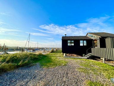 A black wooden house near a marina with boats on water under a blue sky at The Old Fisherman's Hut in Southwold