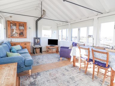 A living room with blue sofa wooden dining table and chairs wood stove and windows at The Old Fisherman s Hut in Southwold