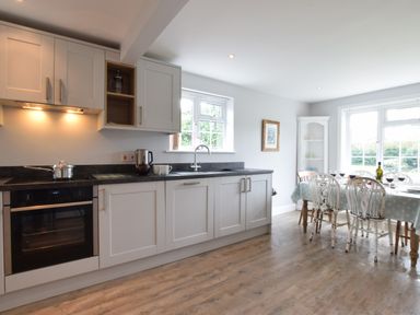 A kitchen with white cabinets, black countertop, an oven, a pot on the stove, a kettle, cups, and a dining table set with glasses and a wine bottle at Hoist Cottage Walberswick