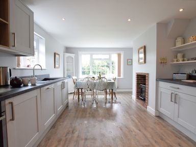 A kitchen with white cabinets a dining table with chairs a window and a fireplace at Hoist Cottage Walberswick