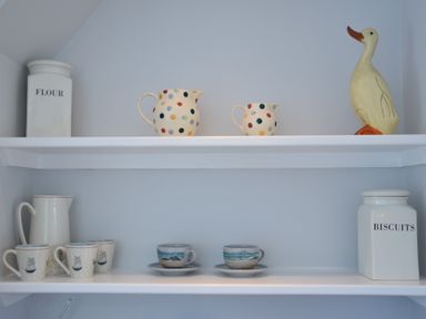 White shelves with flour and biscuits containers ceramic jugs and cups and a duck figurine at Hoist Cottage Walberswick