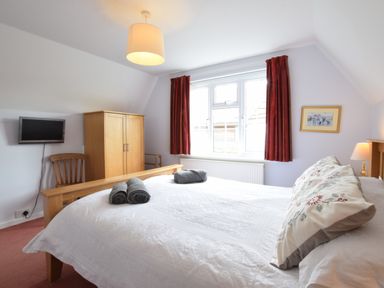 A bedroom with a double bed dressed with pillows and towels a wooden wardrobe a chair a small TV on the wall and a window with red curtains at Hoist Cottage in Walberswick