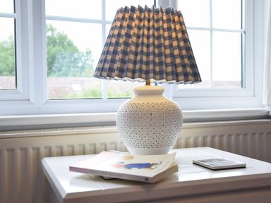 A table with a white ceramic lamp with a checkered shade a book and a coaster near a window at Hoist Cottage Walberswick