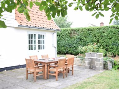 An outdoor patio with a wooden table and six chairs next to a white building with double glass doors at Hoist Cottage Walberswick