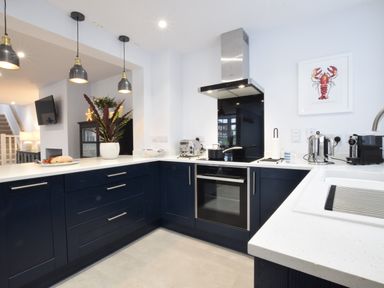 A kitchen with blue cabinets, white countertops, hanging pendant lights, a stove with a pot, a toaster, a kettle, a coffee machine, and a framed lobster picture at Sea Green Cottage Walberswick