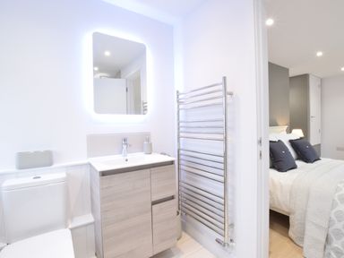 A bathroom with a toilet sink with cabinet a lit mirror and a towel rack next to a bedroom with a bed and pillows at Sea Green Cottage Walberswick